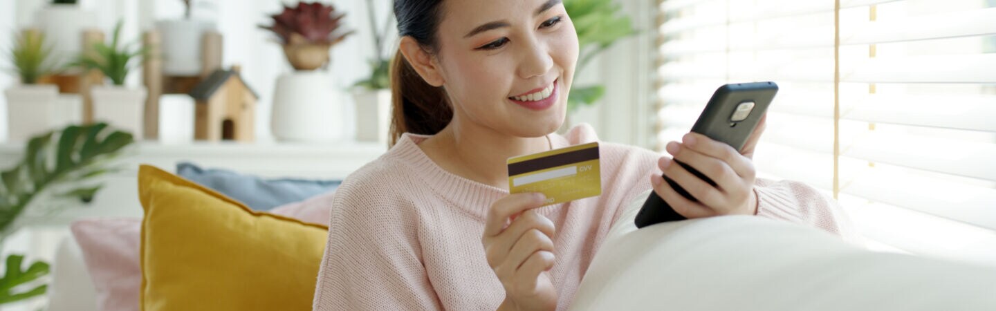 Eine Person sitzt auf einem Sofa, halt eine Bankkarte in der einen Hand und blickt auf ihr Smartphone in der anderen Hand.