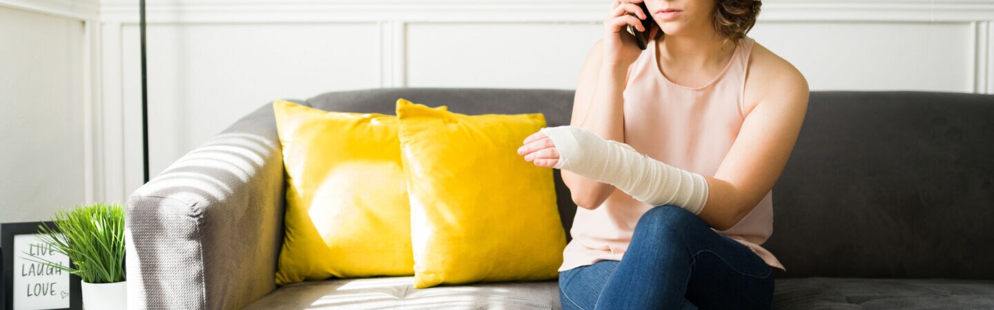 Eine Person sitzt mit einbandagiertem Arm auf einem Sofa und telefoniert.