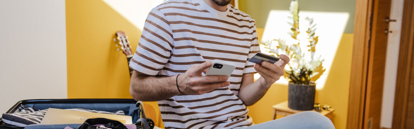 Eine Person sitzt neben einem aufgeklappten Koffer auf dem Bett und hält eine Bankkarte sowie ein Smartphone in den Händen.