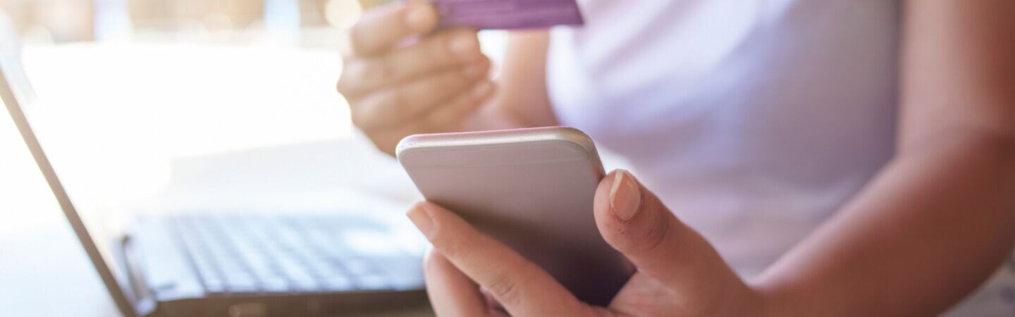 Eine Person sitzt am Tisch vor einem aufgeklappten Notebook. In Händen hält sie Smartphone und Kreditkarte.