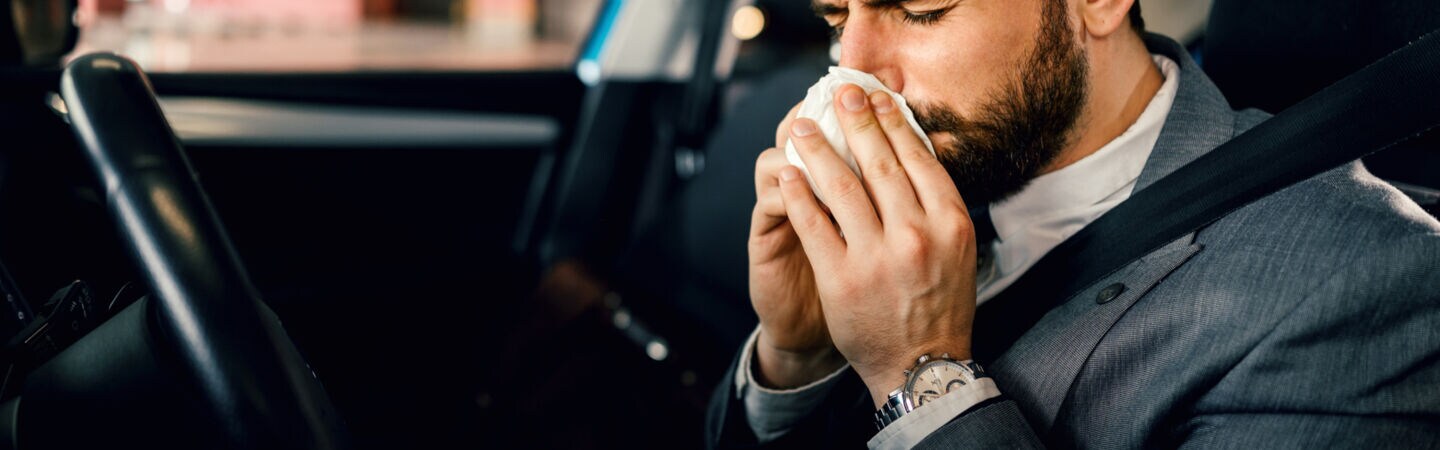 Ein dunkelhaariger Mann im Anzug sitzt angeschnallt am Steuer eines parkenden Autos und putzt seine Nase.