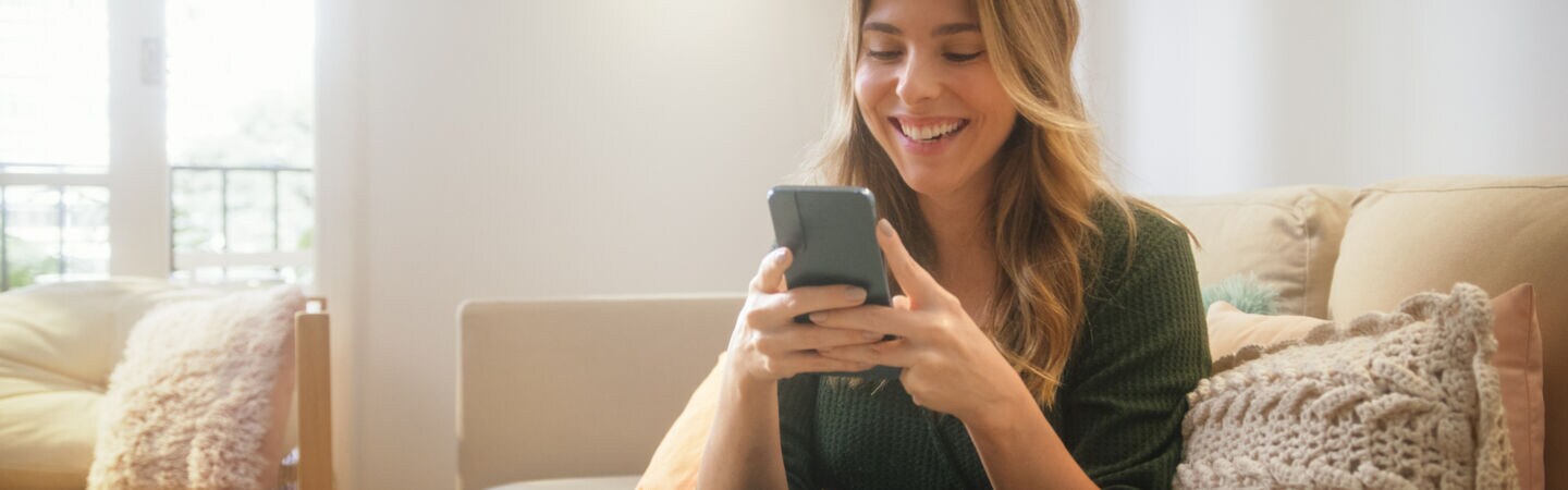 Eine Person sitzt auf einem Sofa und schaut lachend auf das Smartphone, das sie mit beiden Händen festhält.