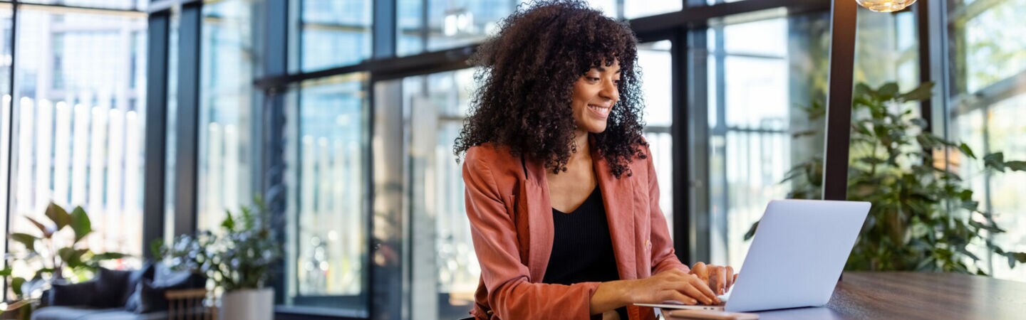 Eine Person sitzt in einem lichtdurchfluteten Ambiente an einem hochbeinigen Tisch und arbeitet am Laptop.