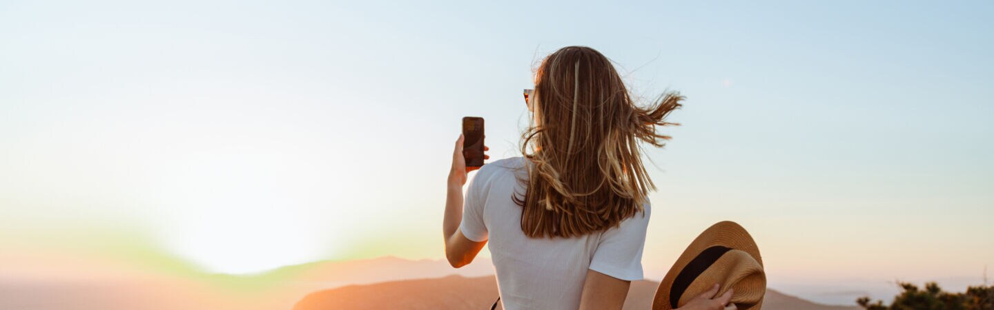 Eine Person in der Rückansicht schaut bei Sonnenuntergang aus dem Dach eines Autos und fotografiert sich dabei.