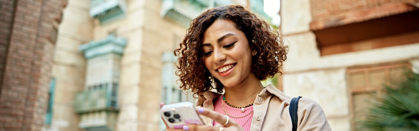 Eine Person steht zwischen hohen Steingebäuden und blickt auf das Smartphone in ihren Händen.