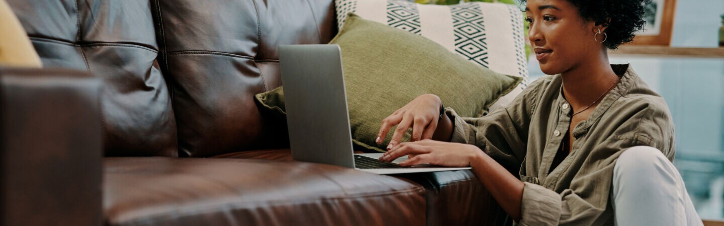 Eine junge Frau sitzt auf dem Boden neben dem Sofa und benutzt ein Notebook.