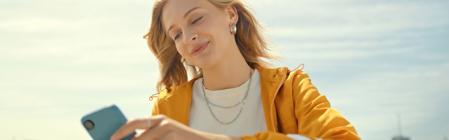 Eine junge Person sitzt im Freien und hält ein Smartphone in ihren Händen.