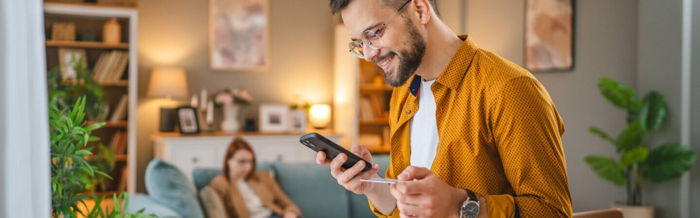 Eine Person sitzt auf einem Tisch und hält eine Kreditkarte sowie ein Smartphone in den Händen. Im Hintergrund sitzt eine weitere Person auf einem Sofa.