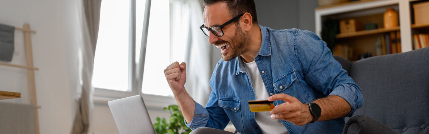 Ein Mann sitzt auf einem Sofa vor einem aufgeklappten Laptop, der auf einem Hocker steht. In einer Hand hält er eine Geldkarte, die andere ballt er zu einer Faust.