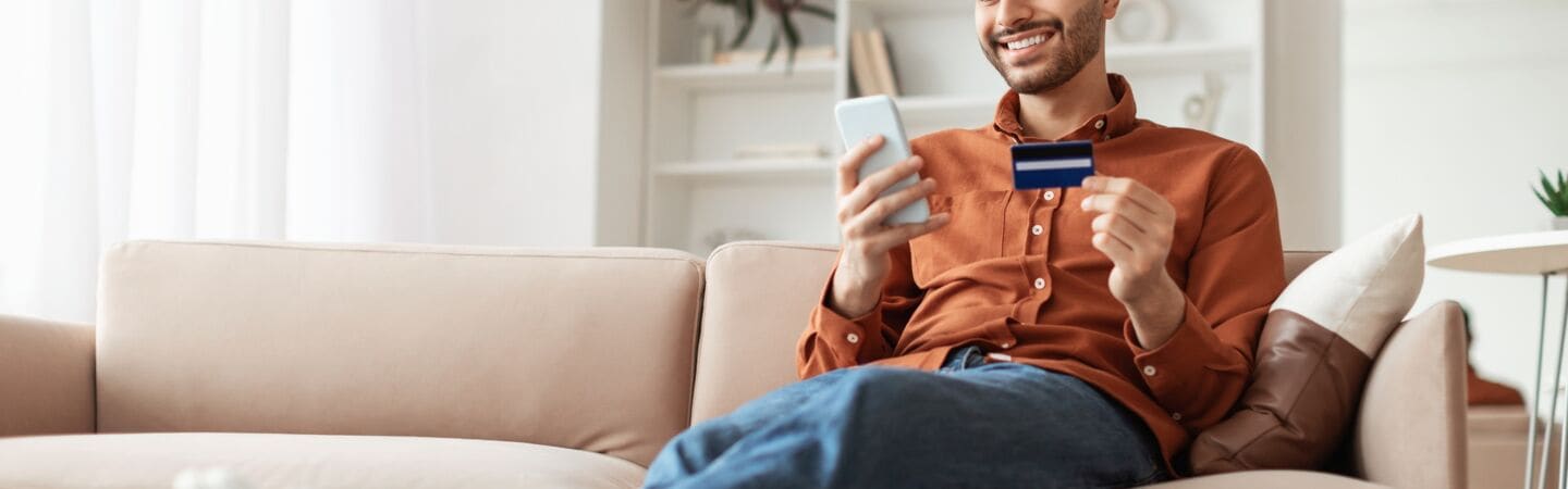 Eine Person sitzt mit ausgestreckten Beinen auf einem Sofa und hält eine Kreditkarte sowie ein Smartphone in den Händen.