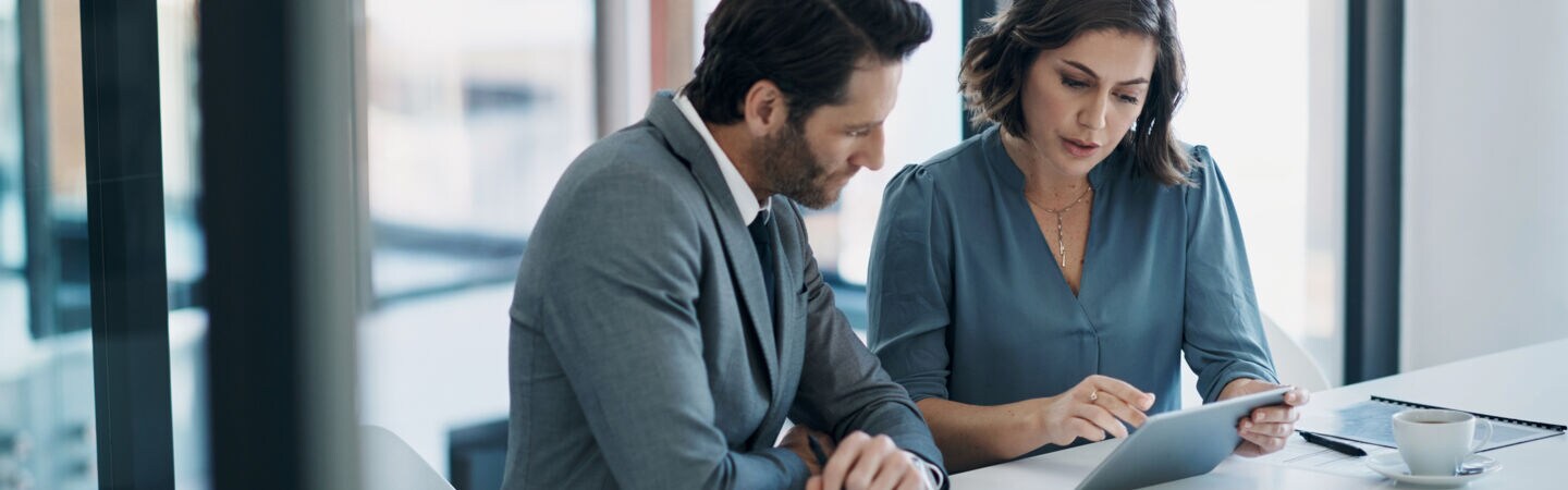 Zwei Personen in Businesskleidung sitzen an einem weißen Tisch, vor sich Kaffeetassen, ein Tablet und diverse Dokumente.