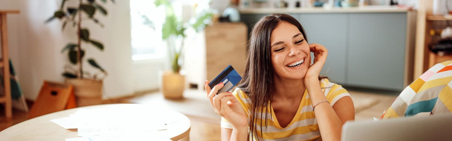 Eine Person sitzt auf dem Fußboden zwischen Couchtisch und Sofa, hält eine Kreditkarte in der Hand und blickt auf ein geöffnetes Notebook, das auf der Sofasitzfläche steht. 