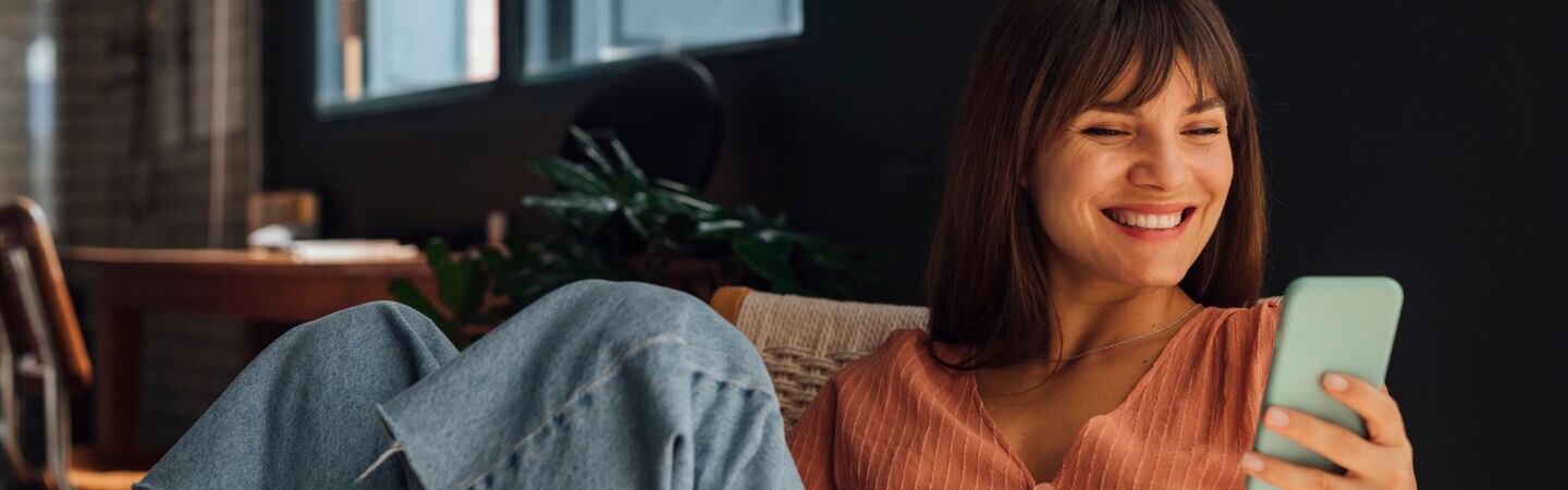 Eine Person sitzt in einem Sessel und blickt lachend auf ihr Smartphone, das sie in der Hand hält.