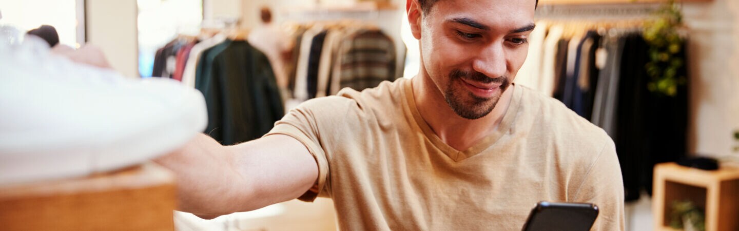 Eine Person steht in einem Bekleidungsgeschäft, greift mit einer Hand nach einem Paar Sneaker und hält in der anderen Hand ein Smartphone.