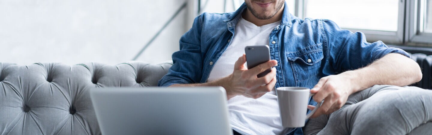 Eine Person sitzt mit ausgestreckten Beinen auf einem Sofa, hat auf dem Schoß ein geöffnetes Laptop liegen und hält in ein Smartphone sowie eine Tasse in den Händen. 