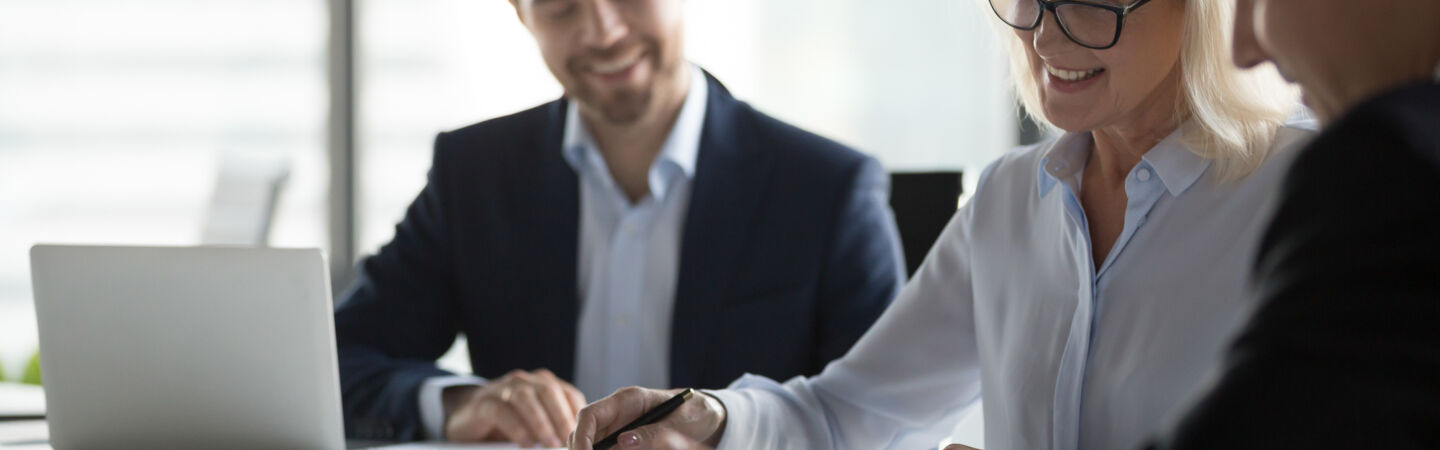 Drei Personen in Businesskleidung sitzen an einem Tisch, auf dem sich ein aufgeklappter Laptop und Papierunterlagen befinden.