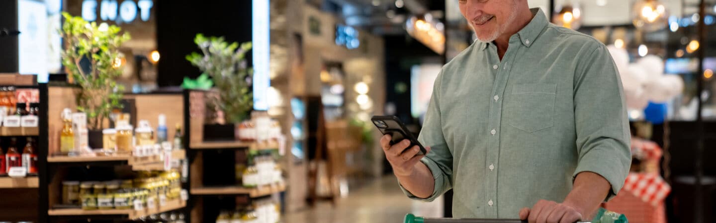 Eine Person steht mit einem Einkaufswagen im Supermarkt und schaut auf ihr Handy in der Hand.
