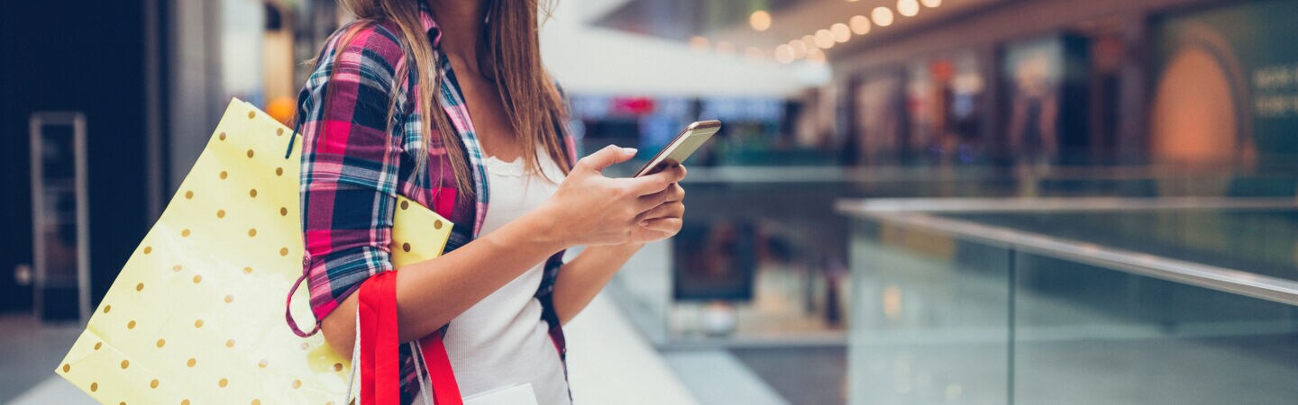 Eine lächelnde Person steht in einer Shoppingmall und hat mehrere Tüten über die Schulter gehängt und hält ein Smartphone in den Händen.