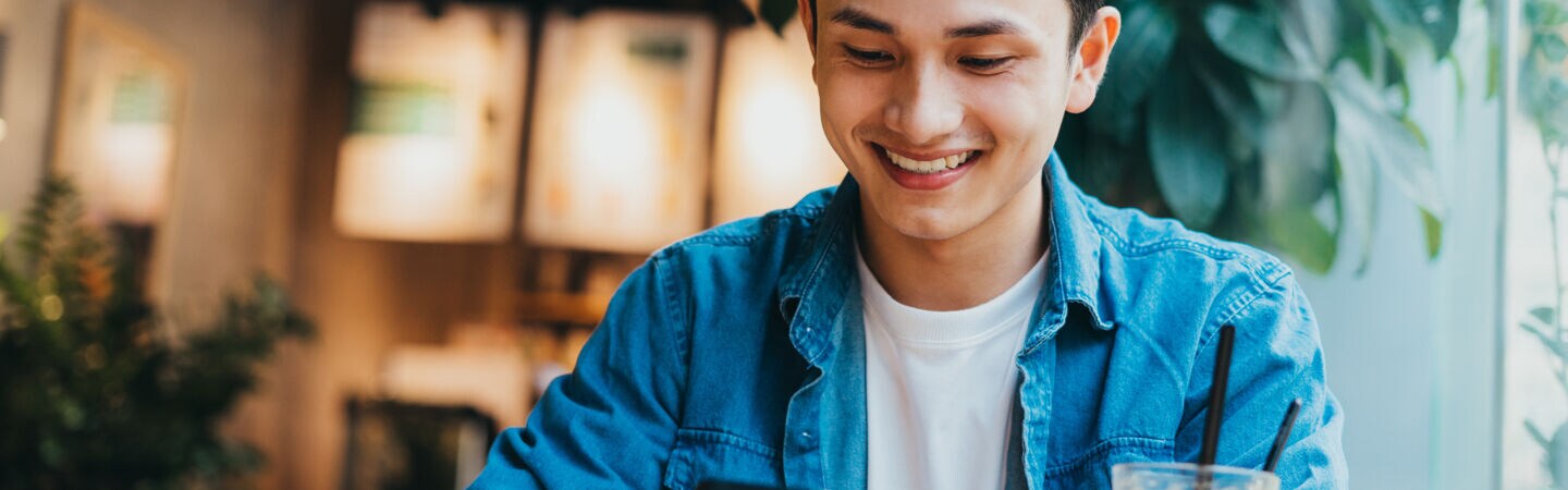 Eine Person sitzt mit einem Café vor sich am Tisch und blickt auf das Smartphone, das sie in den Händen hält.
