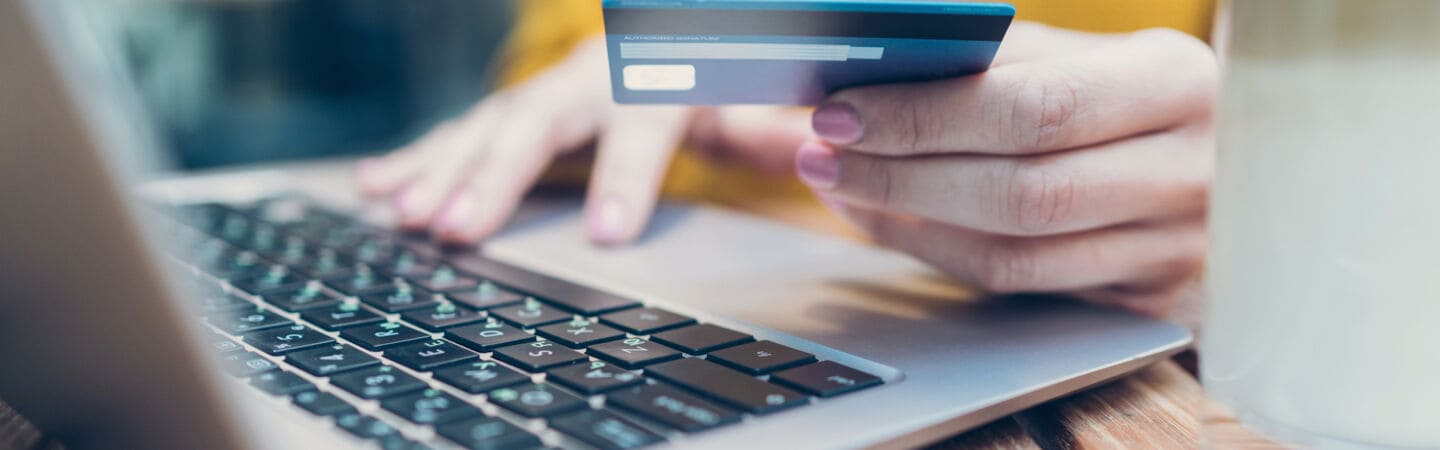 Nahaufnahme einer Person vor einem geöffneten Laptop, in einer Hand eine Bankkarte haltend, die andere liegt auf der Laptoptastatur.