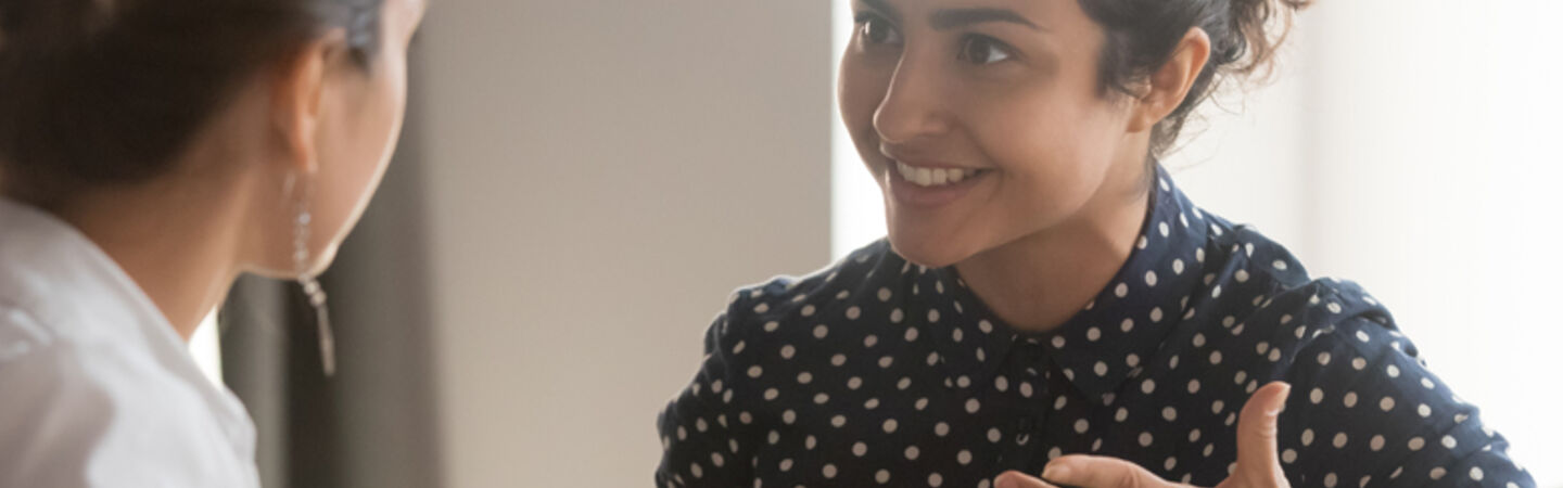 Zwei Mitarbeiterinnen mit verschiedenen kulturellen Hintergründen sitzen an einem Schreibtisch und führen ein Gespräch.