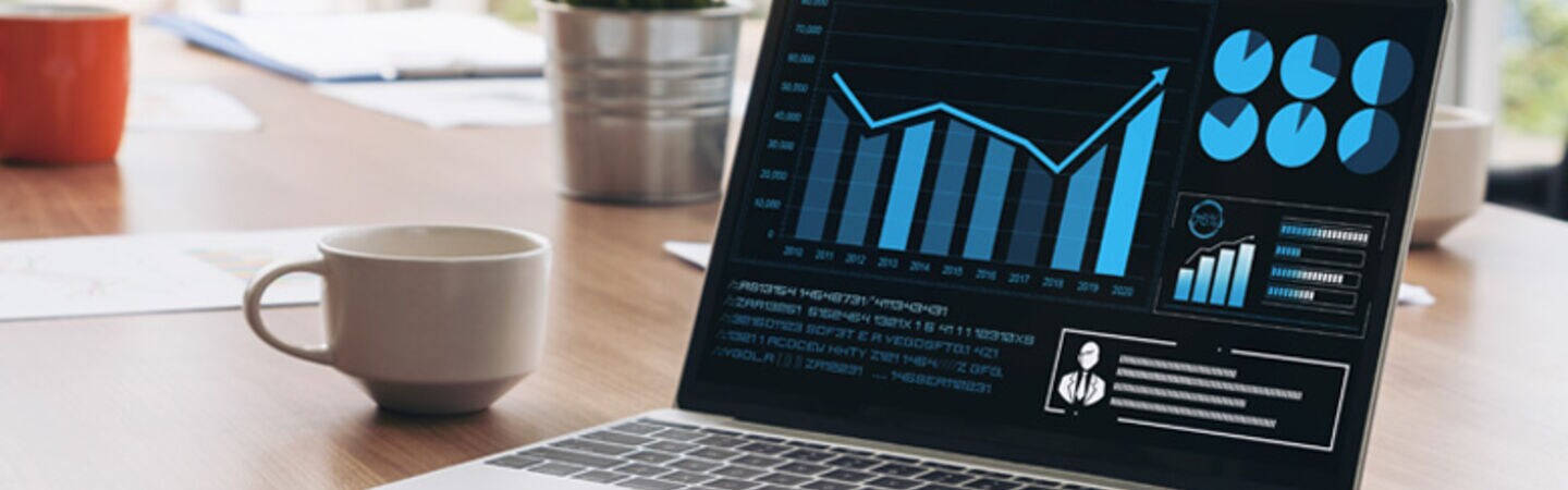 Ein aufgeklappter Laptop, auf dessen Display Diagramme abgebildet sind, steht auf einem Konferenztisch neben einer Kaffeetasse.