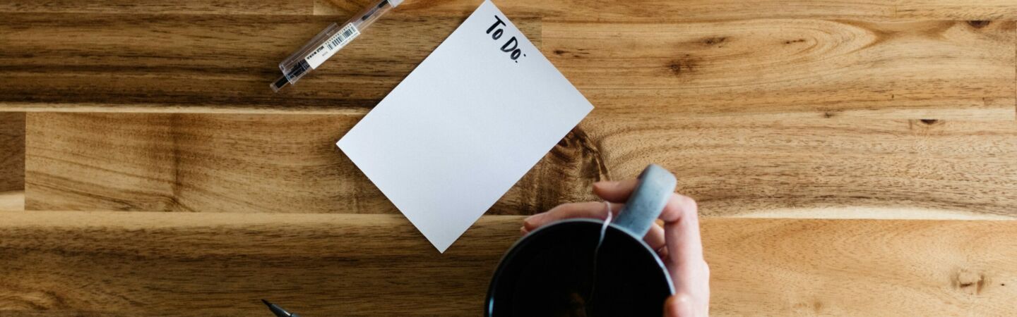 Leere To-do-Liste auf einem Holztisch mit Kaffeetasse und Stift in den Händen einer Person.