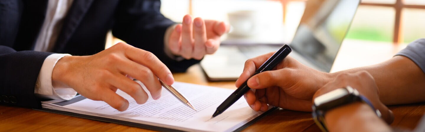 Artikelbild Eine Person im Anzug zeigt auf Dokumente und eine weitere Person unterzeichnet die Dokumente