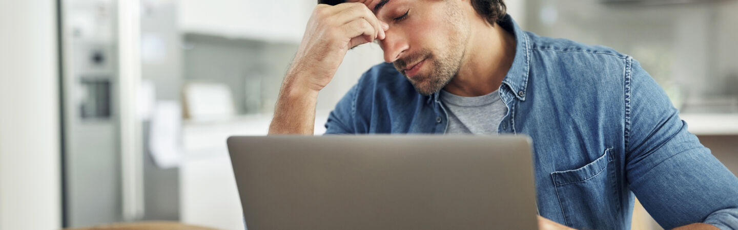 Ein Mann sitzt vor einem Laptop an einem Tisch, hat die Augen geschlossen und eine Hand an seine Stirn gelegt.
