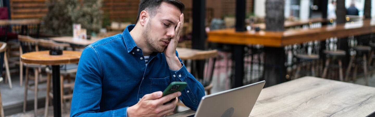 Ein Mann sitzt an einem Tisch vor einem Laptop und hält ein Smartphone in der Hand. Er hat die Augen geschlossen und legt eine Hand über sein Gesicht.