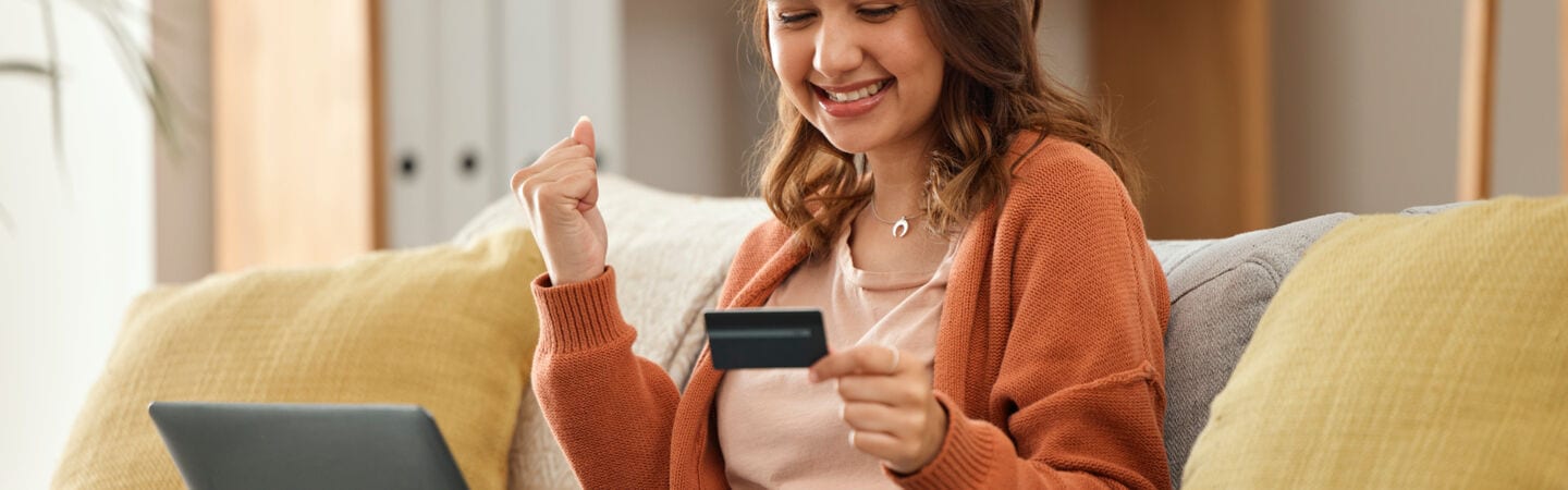 Eine Person sitzt lächelnd auf einem Sofa, einen Laptop auf den Beinen und eine Kreditkarte in der Hand und ballt eine Faust.