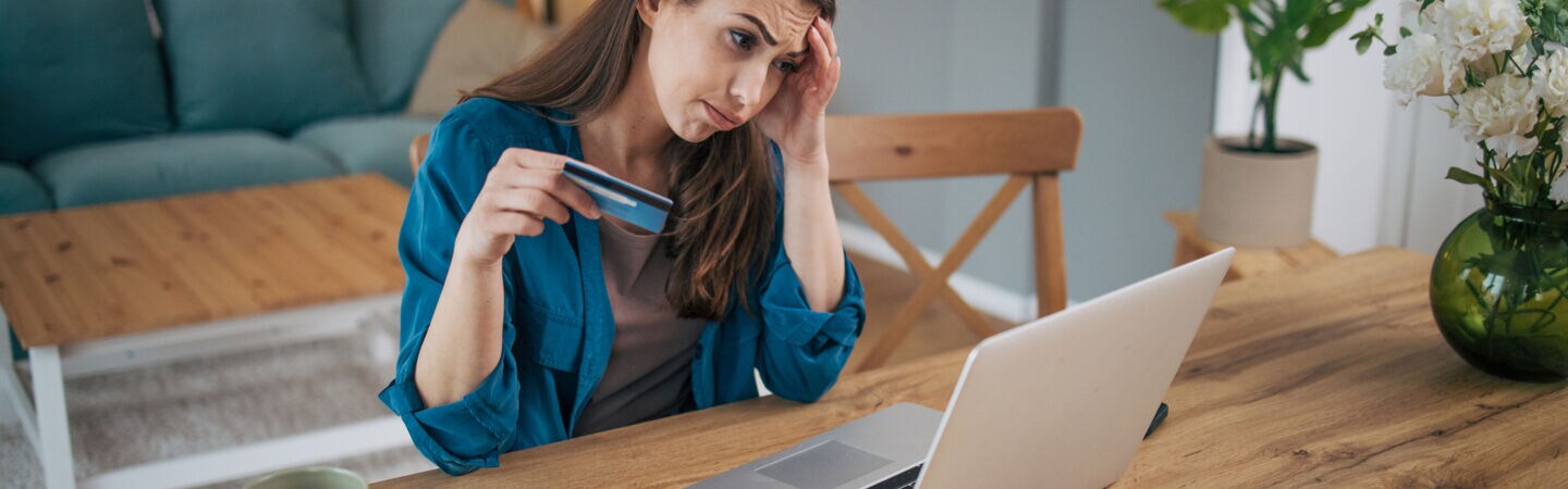 Eine Frau sitzt mit gerunzelter Stirn an einem Tisch vor einem Laptop. In der Hand hält sie eine blaue Karte.