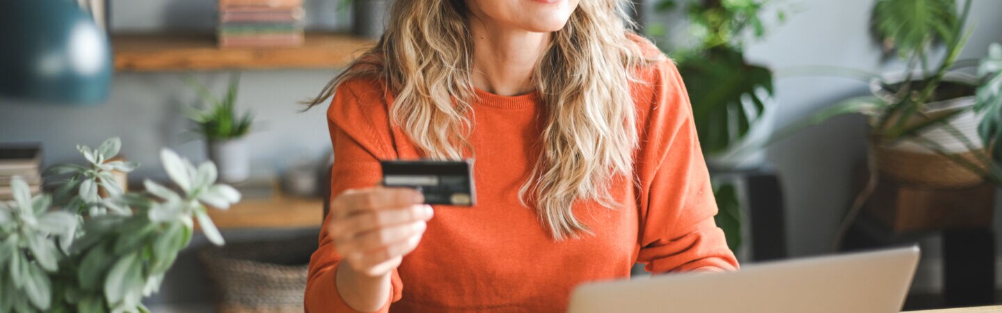 Eine Frau sitzt lächelnd an einem Tisch. Vor ihr steht ein Laptop und in der Hand hält sie eine Kreditkarte.