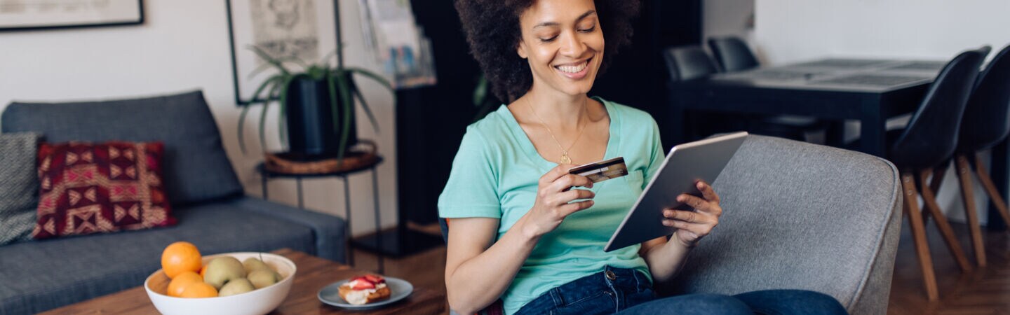 Person sitzt auf einem Sessel und schaut auf ihr Tablet und auf ihre Kreditkarte