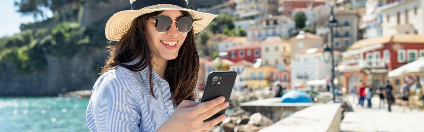 Person schaut auf ihr Handy während sie auf einer Mauer sitzt. Hinter ihr ist das Meer und bunte Häuser