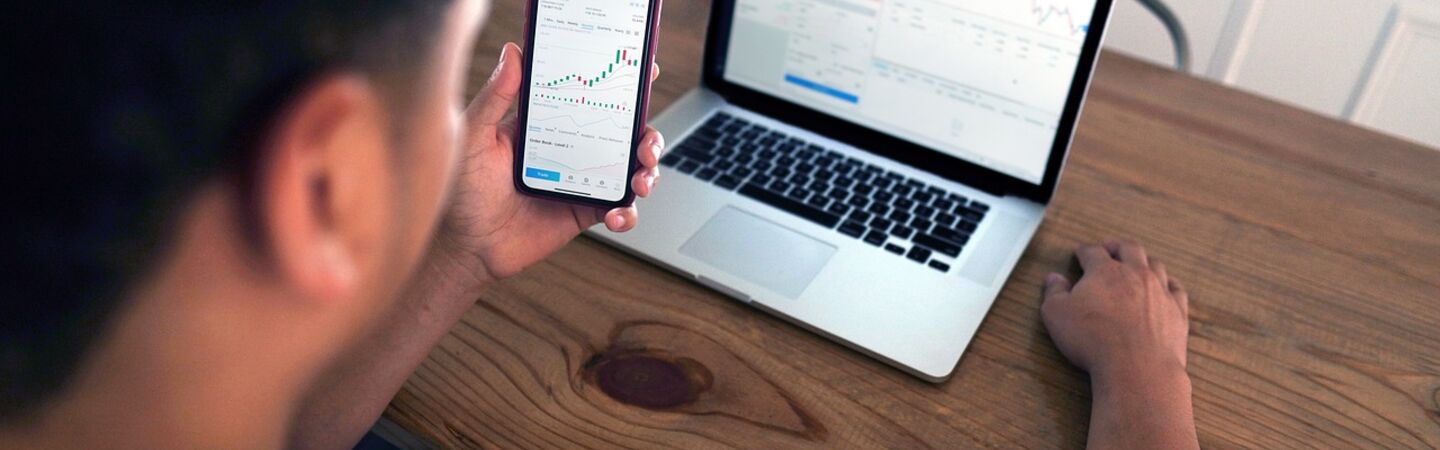 Ein Mann sitzt an einem Holztisch und betrachtet Finanz- und Börsendiagramme auf seinem Smartphone und Laptop. Das Bild zeigt eine moderne Arbeitssituation im Bereich Trading oder Finanzanalyse