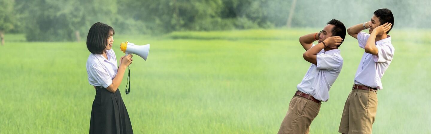 Eine Schülerin steht in einem grünen Feld und spricht durch ein Megafon, während zwei Schüler in Uniform sich die Ohren zuhalten und zurückweichen. Die Szene wirkt humorvoll und zeigt eine lebhafte Kommunikation im Freien