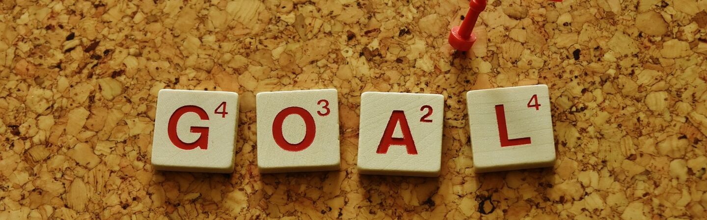 Das Wort ‚GOAL‘ ist aus Scrabble-ähnlichen Buchstabensteinen auf einem Korkbrett gelegt. Daneben steckt eine kleine rote Flagge, die symbolisch ein Ziel oder eine erreichte Etappe markiert.