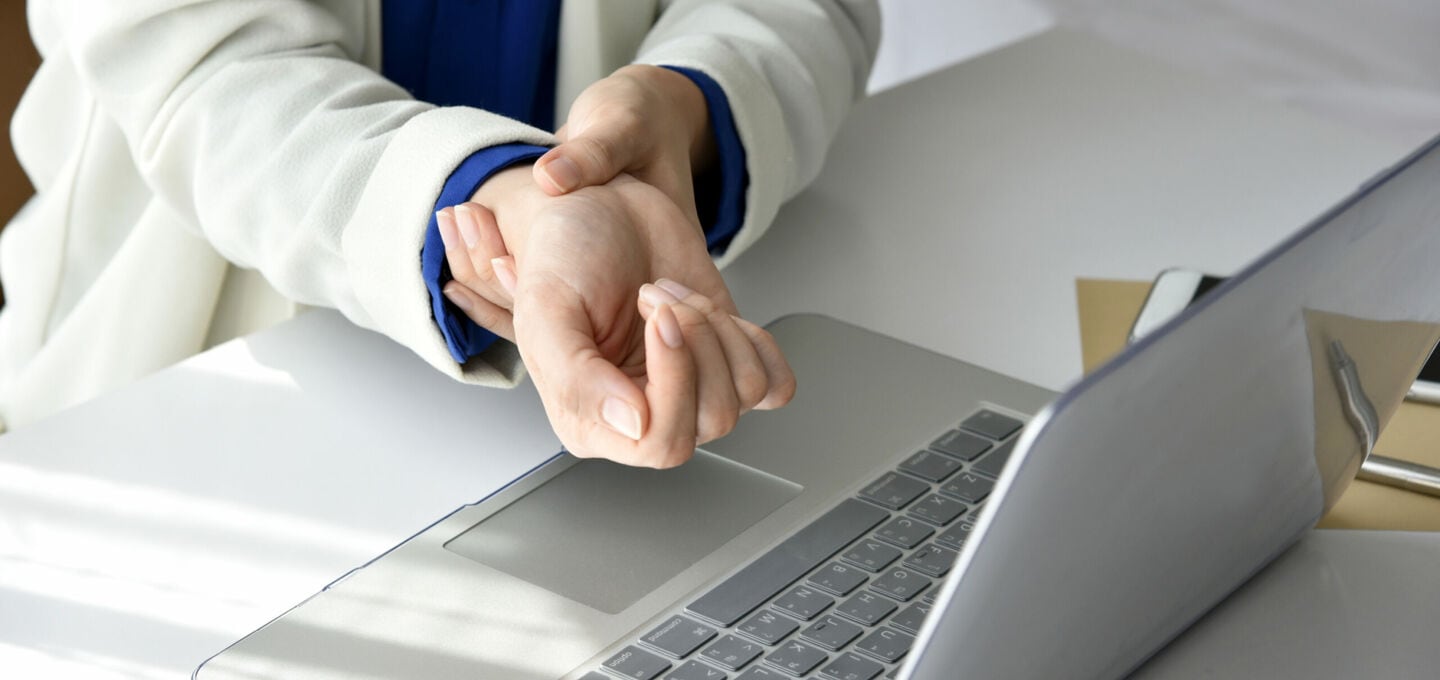 Nahaufnahme einer Person, die am Laptop sitzt und ihr Handgelenk festhält