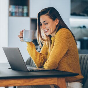 Eine junge Frau sitzt lächelnd mit einer Tasse vor ihrem Laptop