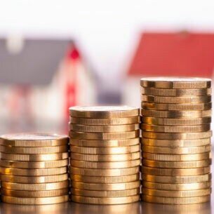 Mehrere Münzstapel aneinandergereiht während im Hintergrund kleine Modellhäuser stehen.