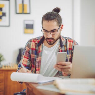Ein junger Mann an einem Tisch beim Notizenmachen mit Smartphone in einer Hand