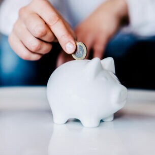 Nahaufnahme der Hand einer Person, die eine Münze in ein weißes Sparschwein wirft