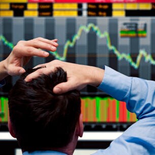 Ein Bildschirm zeigt fallende Börsenkurse und eine Person schlägt die Hände über dem Kopf zusammen.