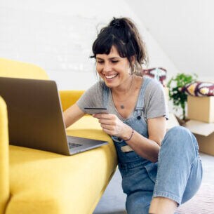Eine junge Frau mit Kreditkarte in einer Hand vor einem gelben Sofa, auf dem ein Laptop steht