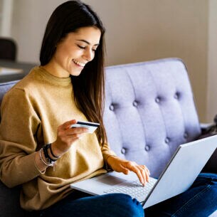 Eine Frau im Schneidersitz auf einem Sofa mit Laptop im Schoß und Kreditkarte in der Hand
