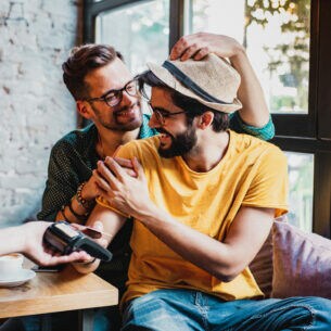Zwei verliebte Männer bezahlen lächelnd und kontaktlos in einer Bar.