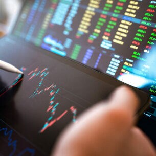 Eine Hand mit einem Stift, der auf ein Tablet mit Börsenkursen tippt, dahinter ein Display mit weiteren Zahlen