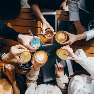 Table-top-Ansicht einer Gruppe von jungen Menschen, die mit verschiedenen Getränken in der Hand anstoßen. Auf dem Tisch darunter liegt ein Tablet, ein Smartphone und Dokumente.