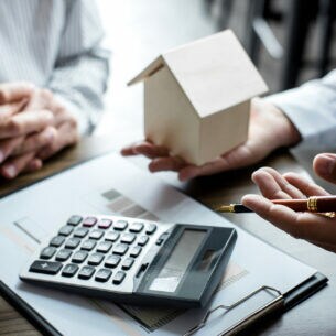 Zwei Personen, eine mit Modellhaus in der Hand, an einem Tisch mit Taschenrechner und Clipboard